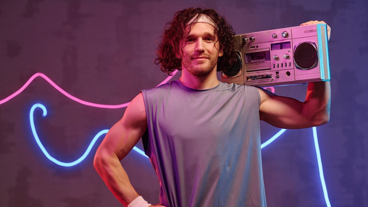 A curly-haired man in a sleeveless gray tee and headband holds a silver boombox on his shoulder in front of pink and blue neon lights.