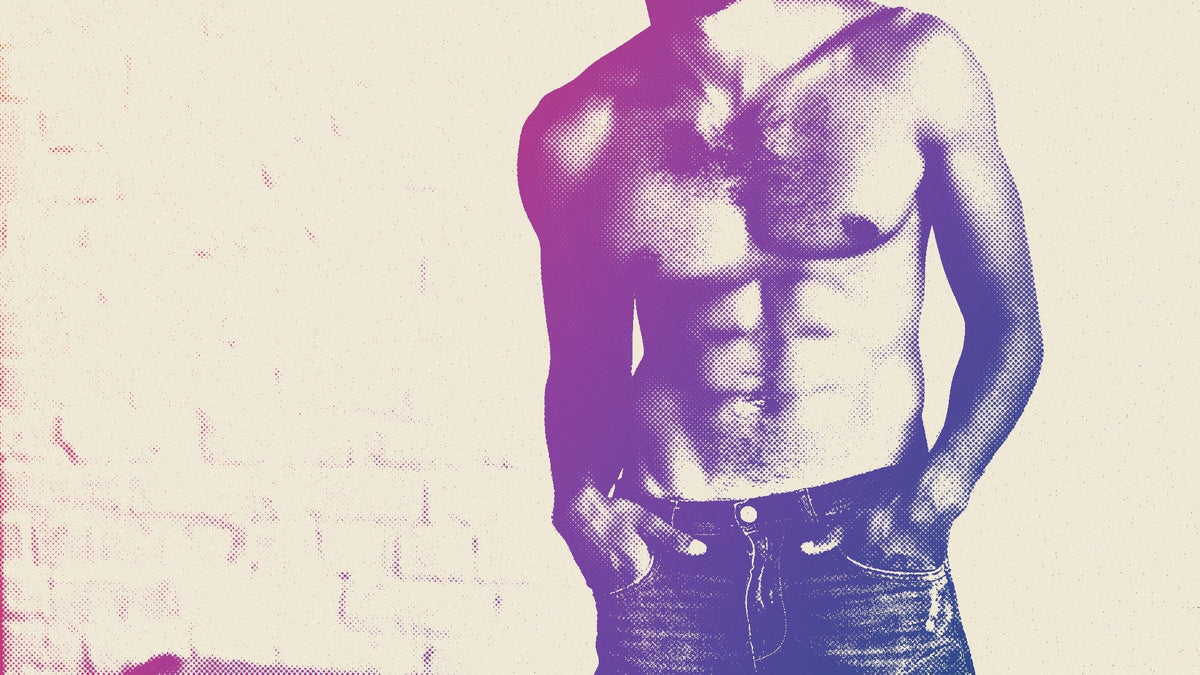 A shirtless, muscular man with a hairy chest stands with his hands in the pockets of his blue jeans against a white brick wall, cropped at the jaw.