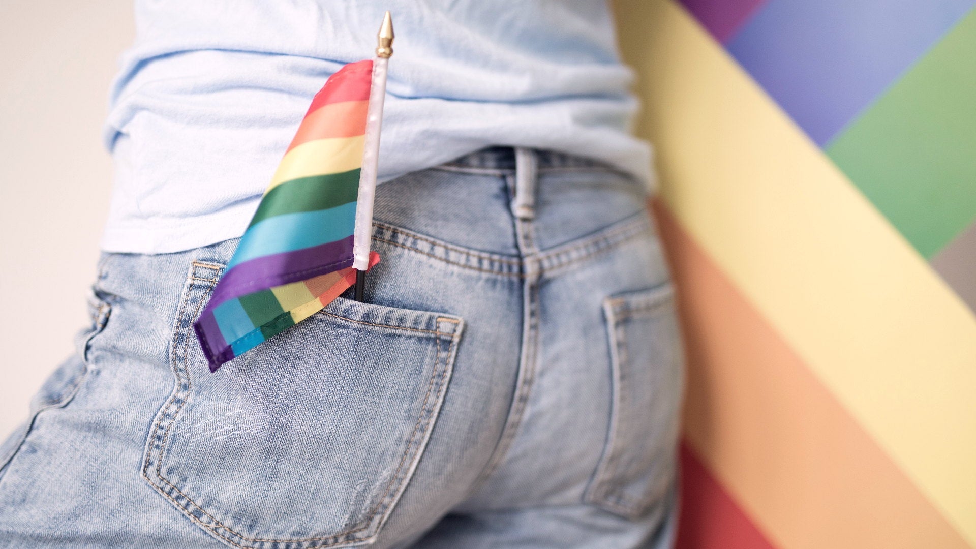 A rainbow pride flag tucked into the back pocket of light blue jeans, a large rainbow flag blurred in the background.