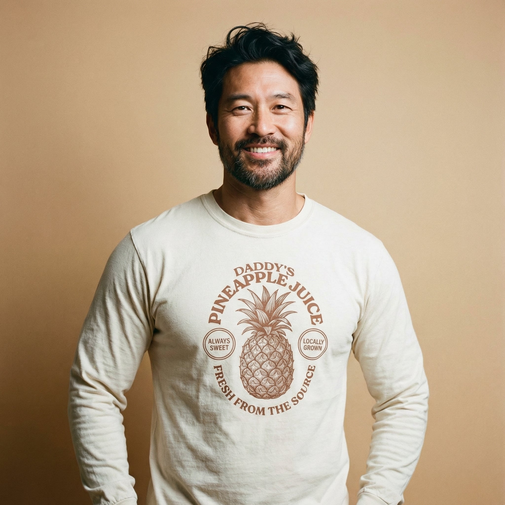 A front-facing portrait of a smiling middle-aged man with dark, slightly messy hair and a salt-and-pepper beard against a solid tan background. He is wearing a cream-colored long-sleeved t-shirt with a brown vintage-style graphic centered on the chest. The graphic depicts a detailed line-art pineapple surrounded by circular text that reads 'Daddy's Pineapple Juice' at the top, 'Always Sweet' and 'Locally Grown' in small circles on the sides, and 'Fresh From The Source' along the bottom edge. #color_Ivory