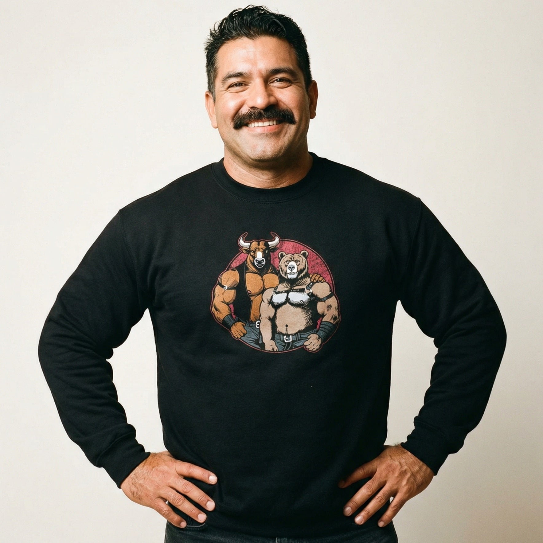 A portrait of a middle-aged man with short dark hair and a full mustache, smiling warmly at the camera with his hands on his hips. He is wearing a black crewneck sweatshirt that features a circular graphic in the center. Inside the red-toned circle, there are two muscular, anthropomorphic animals—a bull and a bear—styled in leather subculture gear, including harnesses and vests. The bull has its arm around the bear's shoulder. The background is a plain, light-colored wall. #color_Black