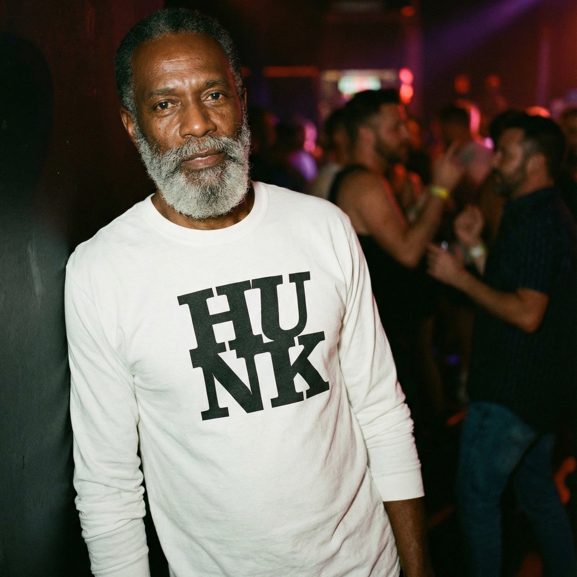 A portrait of an older Black man with a full gray beard leaning against a dark wall in a nightclub. He is wearing a white long-sleeved shirt with the word 'HUNK' printed in large, black block letters across the chest. The background is softly blurred, showing other patrons socializing under dim, colorful club lighting, creating a relaxed and cool atmosphere. #color_White