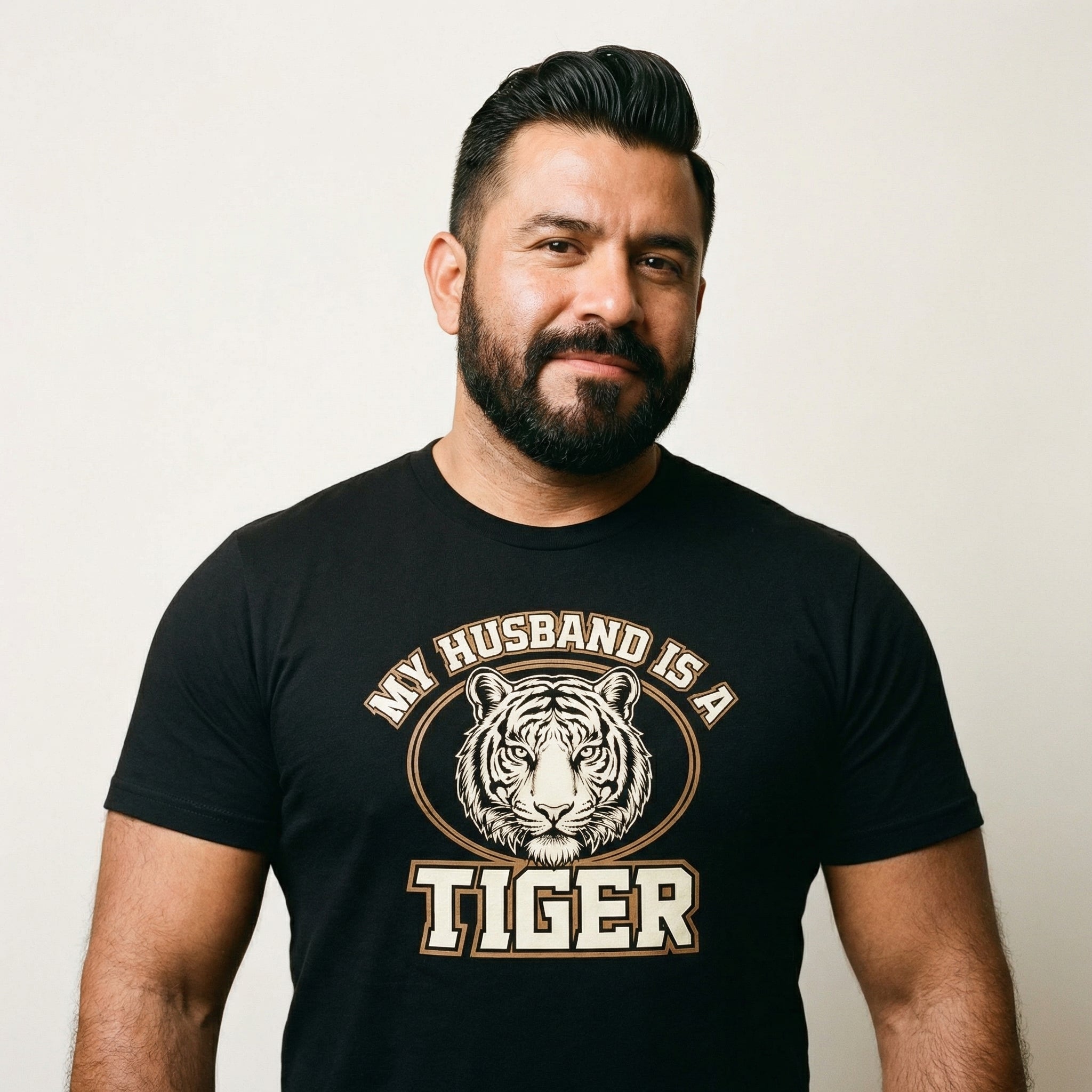 A waist-up portrait of a man with dark hair and a full beard, smiling slightly at the camera. He is wearing a black t-shirt featuring a graphic of a tiger's face inside an oval frame with the text 'MY HUSBAND IS A TIGER' printed around it. The background is a plain, light-colored wall.