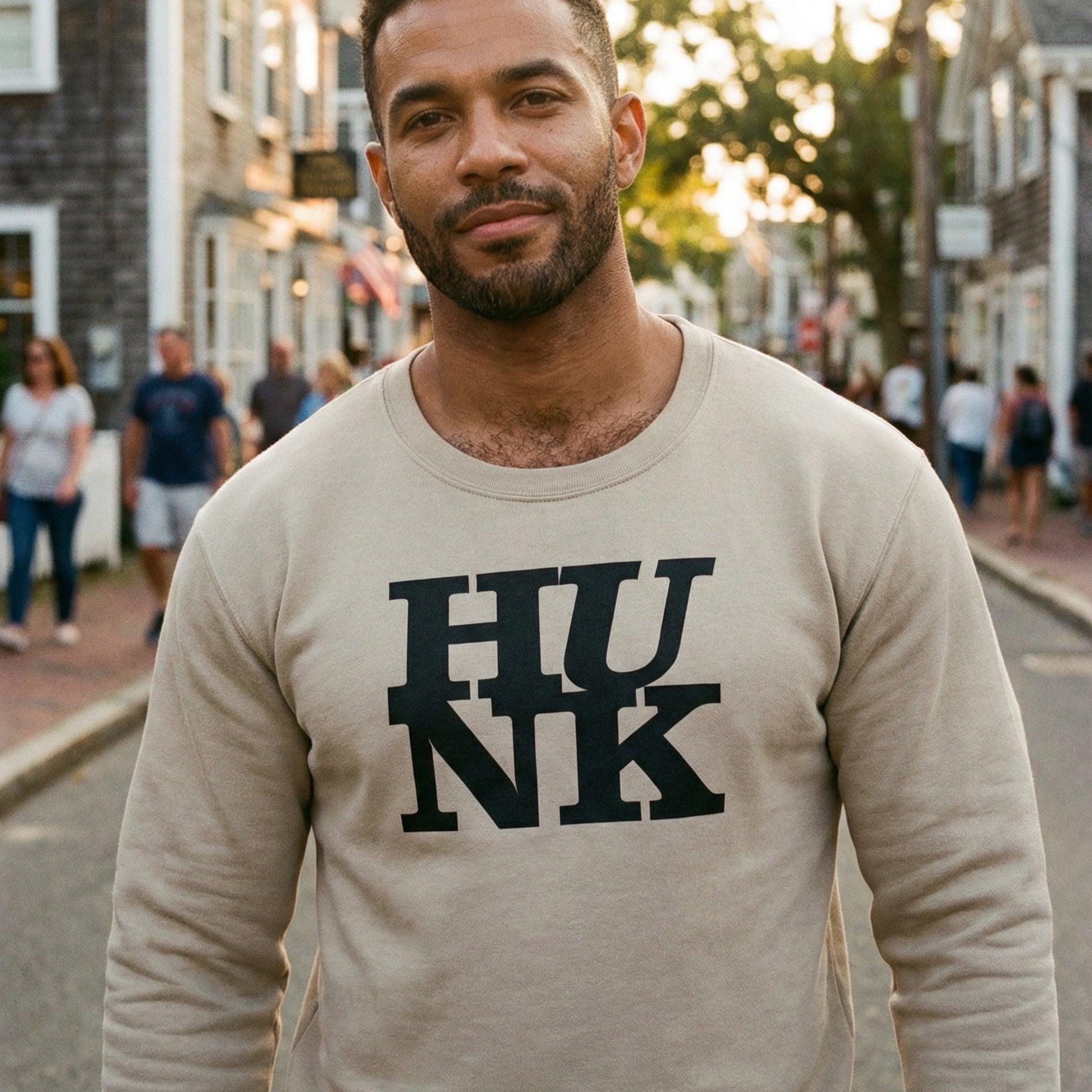 A medium close-up of a man with short dark hair and a beard, looking calmly at the camera with a subtle smile. He is wearing a tan sweatshirt featuring the word "HUNK" in bold, black, blocky letters stacked in two rows. The background shows a charming, sun-drenched street in a coastal town with grey-shingled buildings and people walking in the distance. #color_Sand