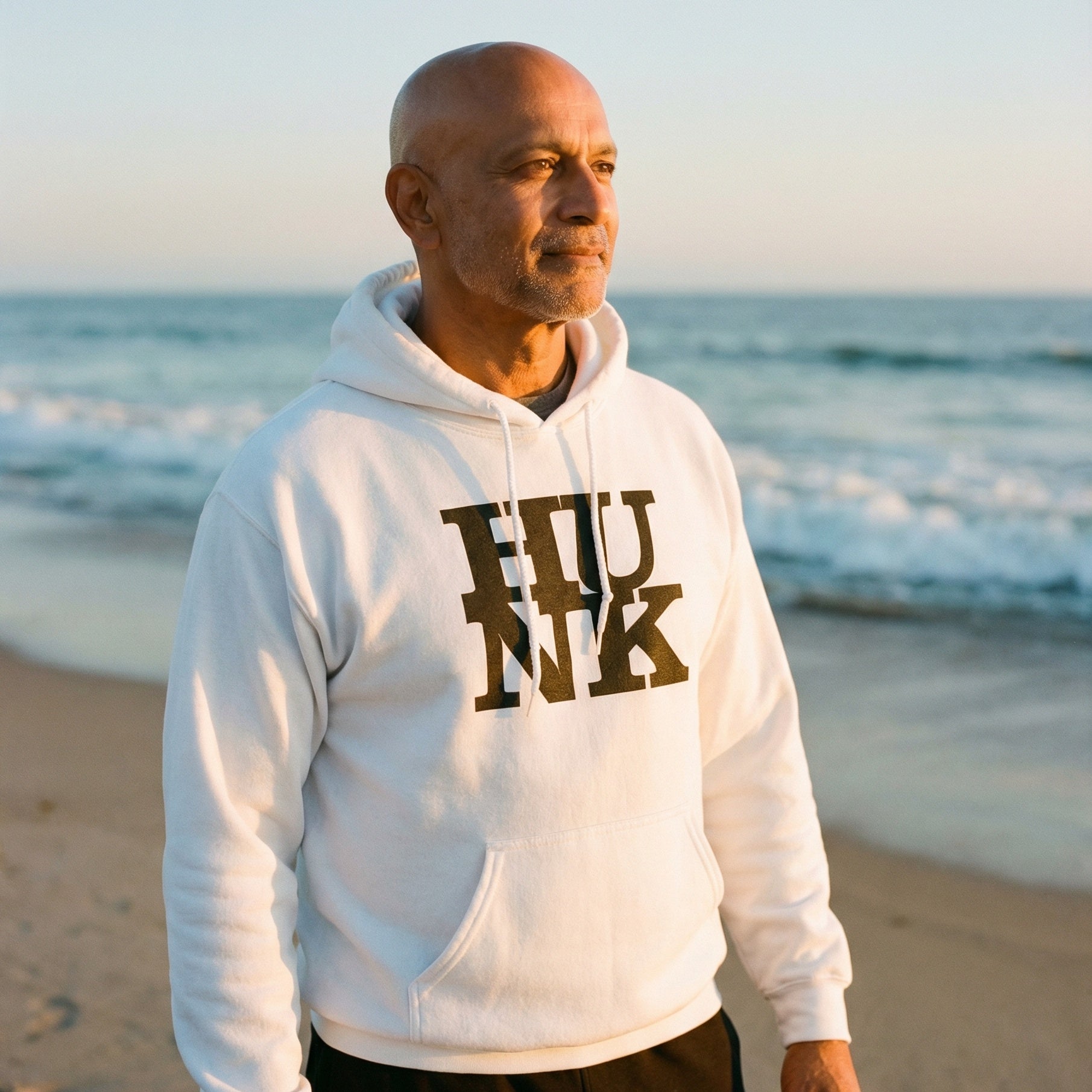 A medium shot of a bald, middle-aged man with a graying beard, looking thoughtfully toward the horizon. He is wearing a white pullover hoodie with the word "HUNK" printed in large, black, stylized block letters on the chest. The background shows a peaceful beach setting with gentle waves rolling onto the sand under the soft, warm light of a setting or rising sun. The overall atmosphere is calm and reflective. #color_White