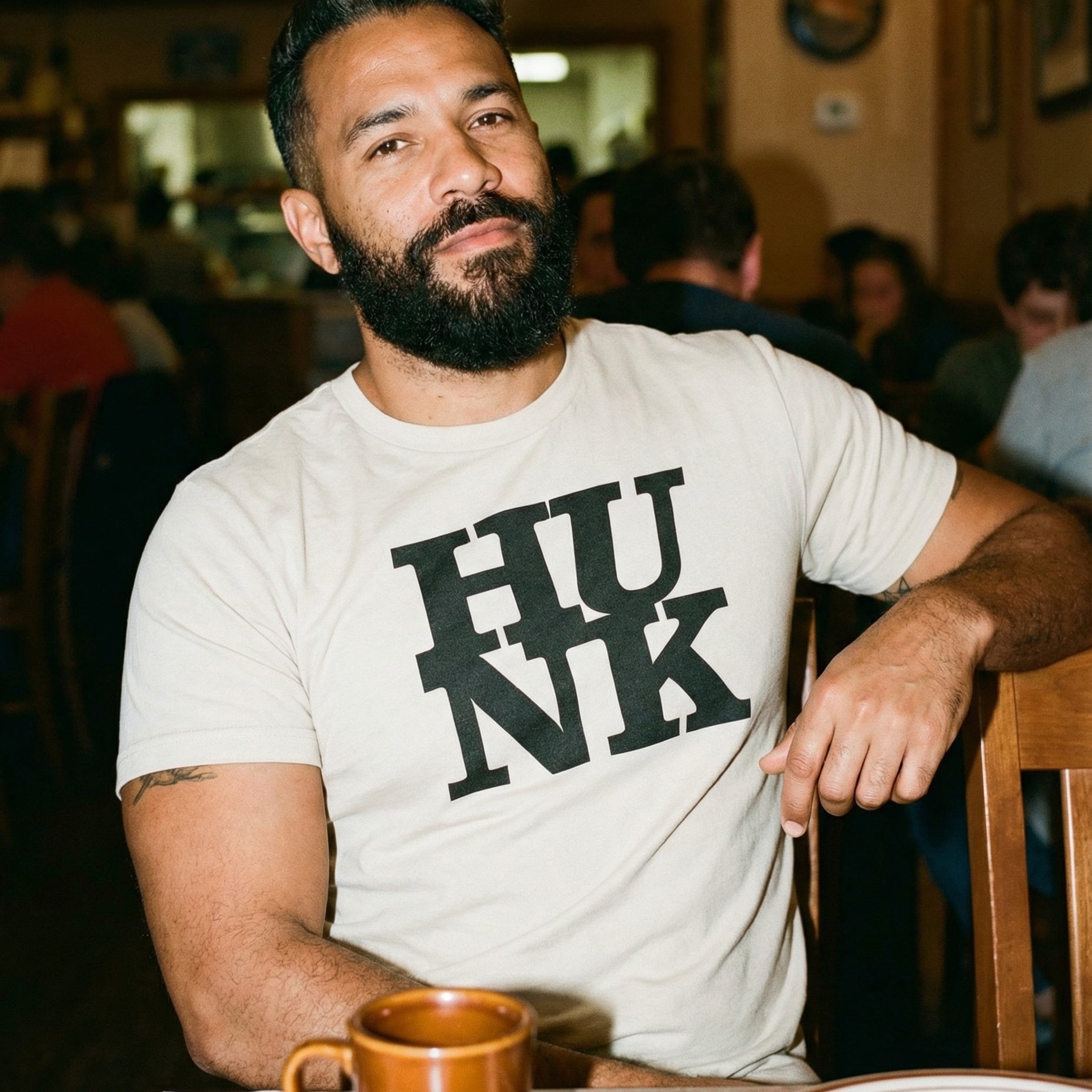 A close-up portrait of a man with a thick black beard and short hair, wearing a cream-colored t-shirt with the word 'HUNK' printed in bold black serif letters. He is sitting at a wooden table in a dimly lit diner, looking directly at the camera with a slight smile. A brown ceramic mug sits on the table in front of him, and other patrons are blurred in the background. #color_Vintage White