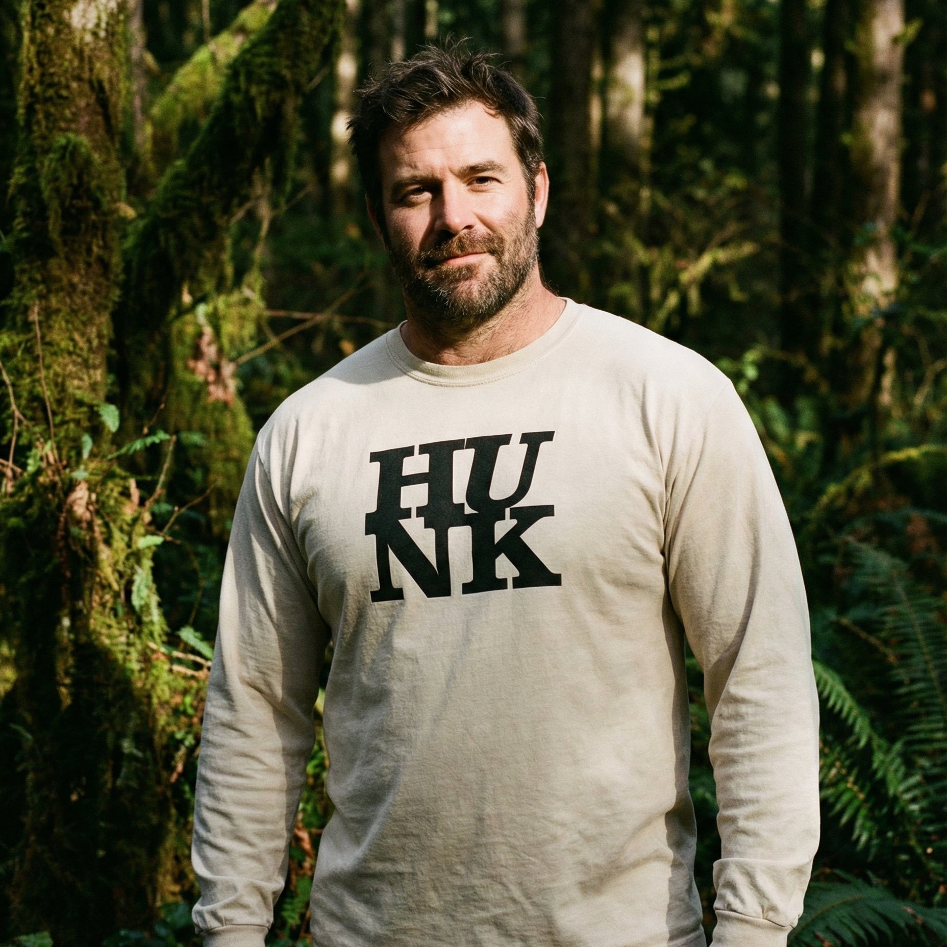 A medium shot of a man with short brown hair and a beard, smiling slightly while standing in a dense, mossy forest. He is wearing a beige long-sleeved t-shirt with 'HUNK' printed in bold, black, stylized letters on the chest. The background is filled with green, moss-covered tree trunks and ferns, with soft sunlight filtering through the canopy. #color_Ivory