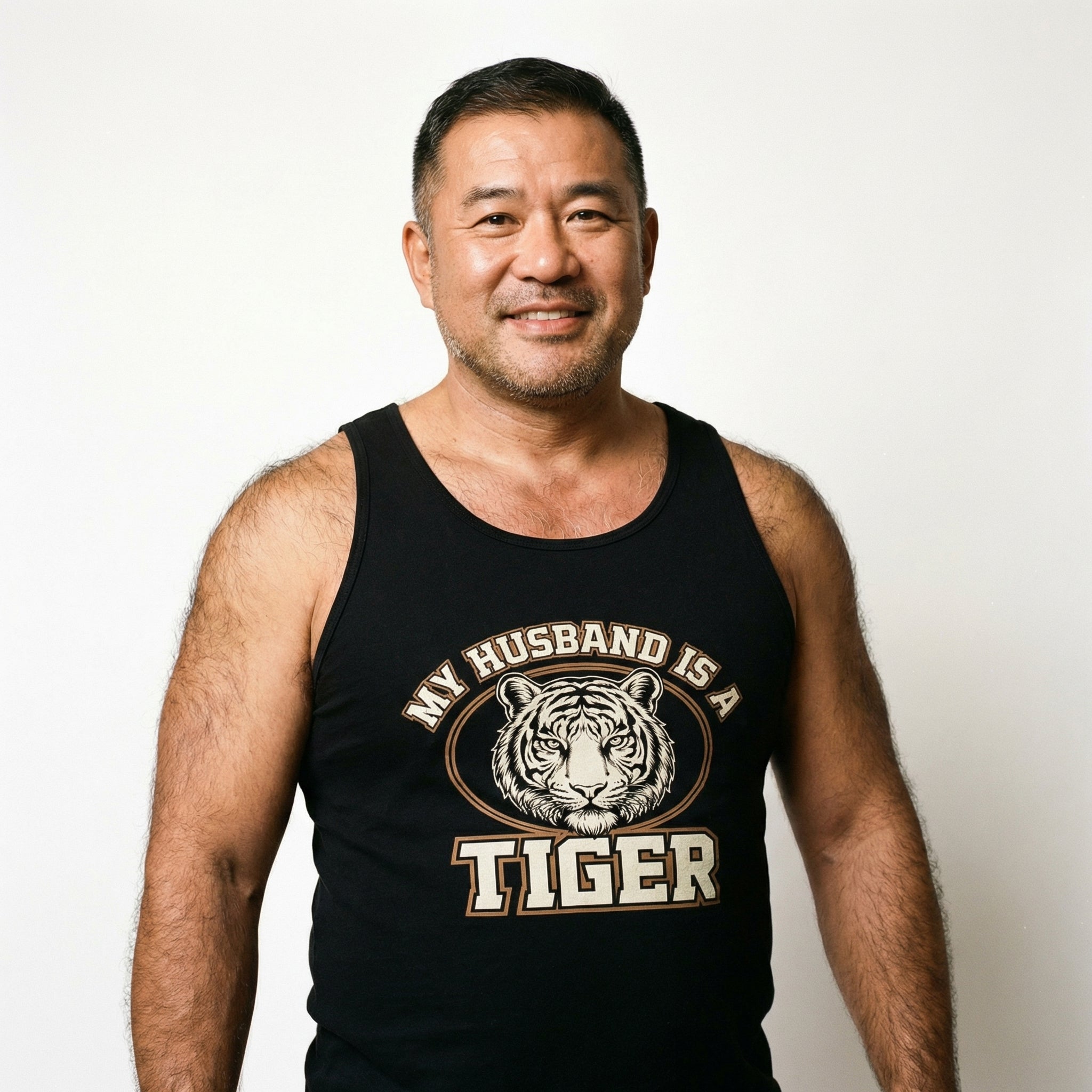 A medium shot of a smiling middle-aged East Asian man with short dark hair and light stubble. He is standing against a plain white background, wearing a black tank top that features a graphic of a tiger's face and the text "MY HUSBAND IS A TIGER." The man has a friendly expression and visible hair on his muscular arms and shoulders. #color_Black