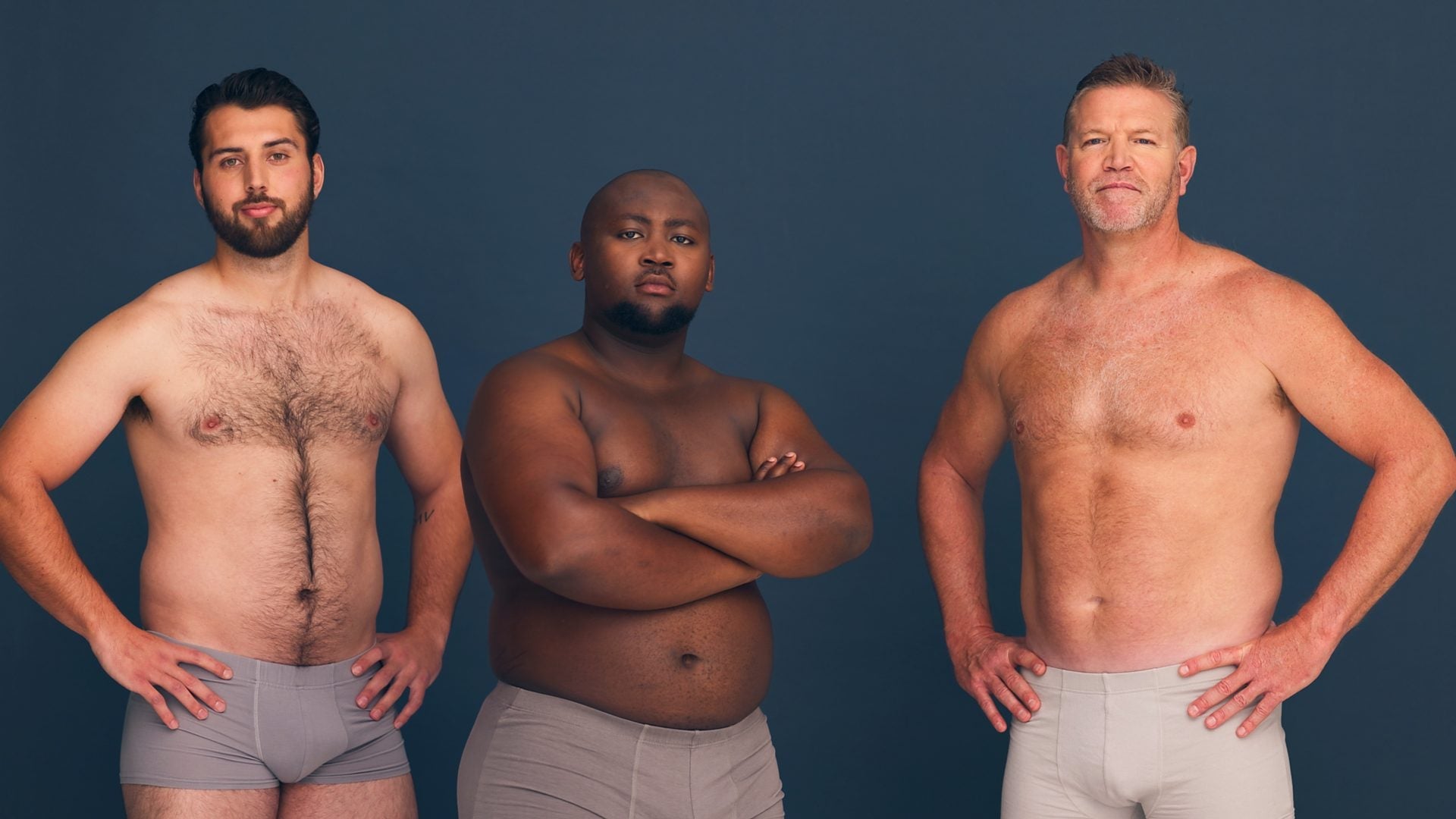 Three men of different builds and body hair standing side by side against a dark blue backdrop.