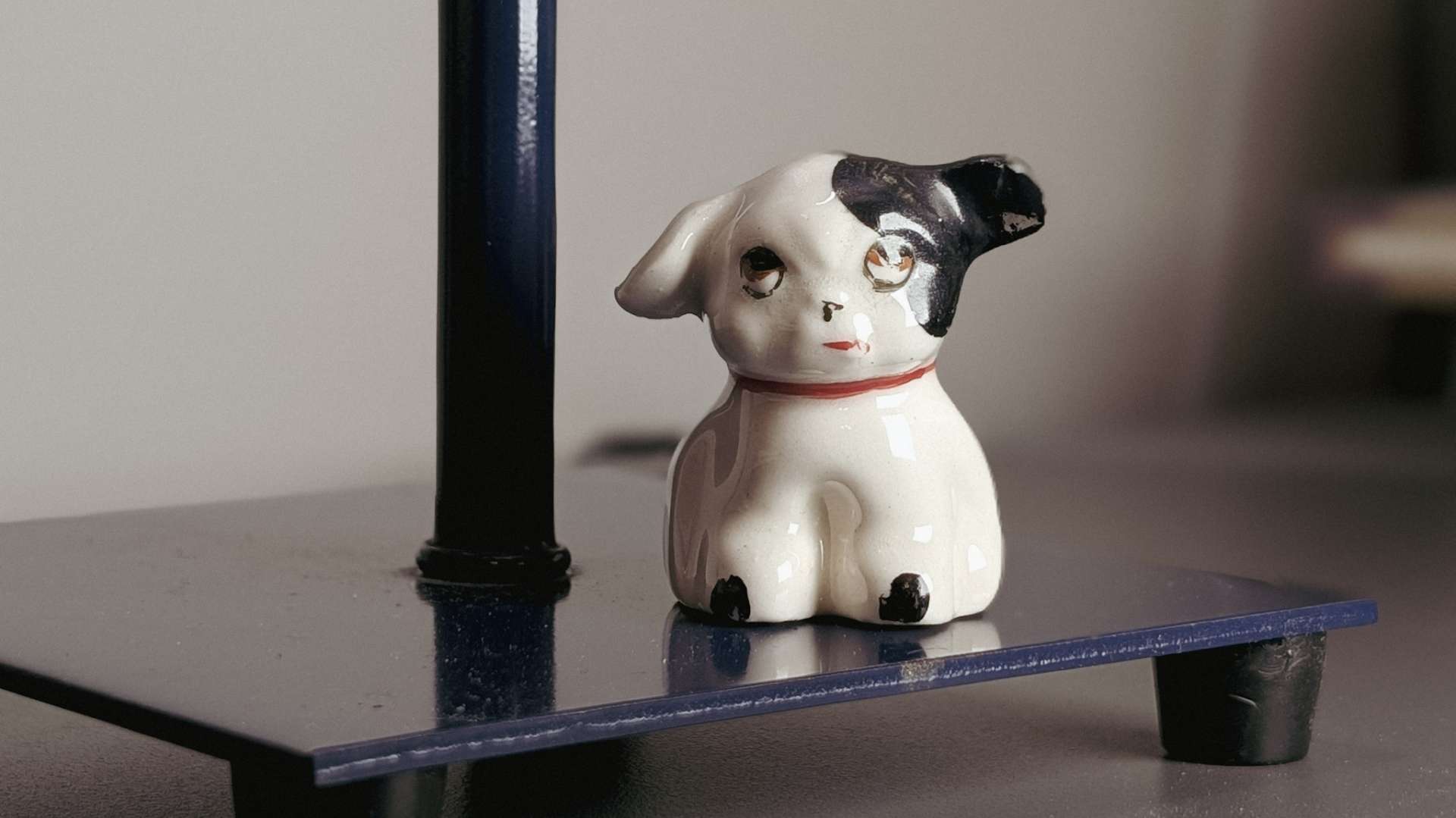 Small vintage porcelain dog figurine, white with a black patch over one eye and ear and a painted red collar, sitting on a navy lamp base.