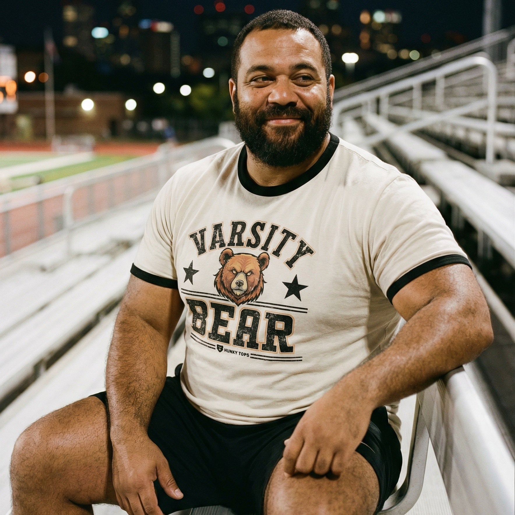 A man with a short, dark beard and a warm smile is seated on outdoor metal stadium bleachers. He wears a cream-colored ringer t-shirt with black trim around the neck and sleeves, featuring a graphic that says 'VARSITY BEAR' with a bear's face. The background shows a blurred sports field and city lights under a dark night sky. #color_Natural/Black