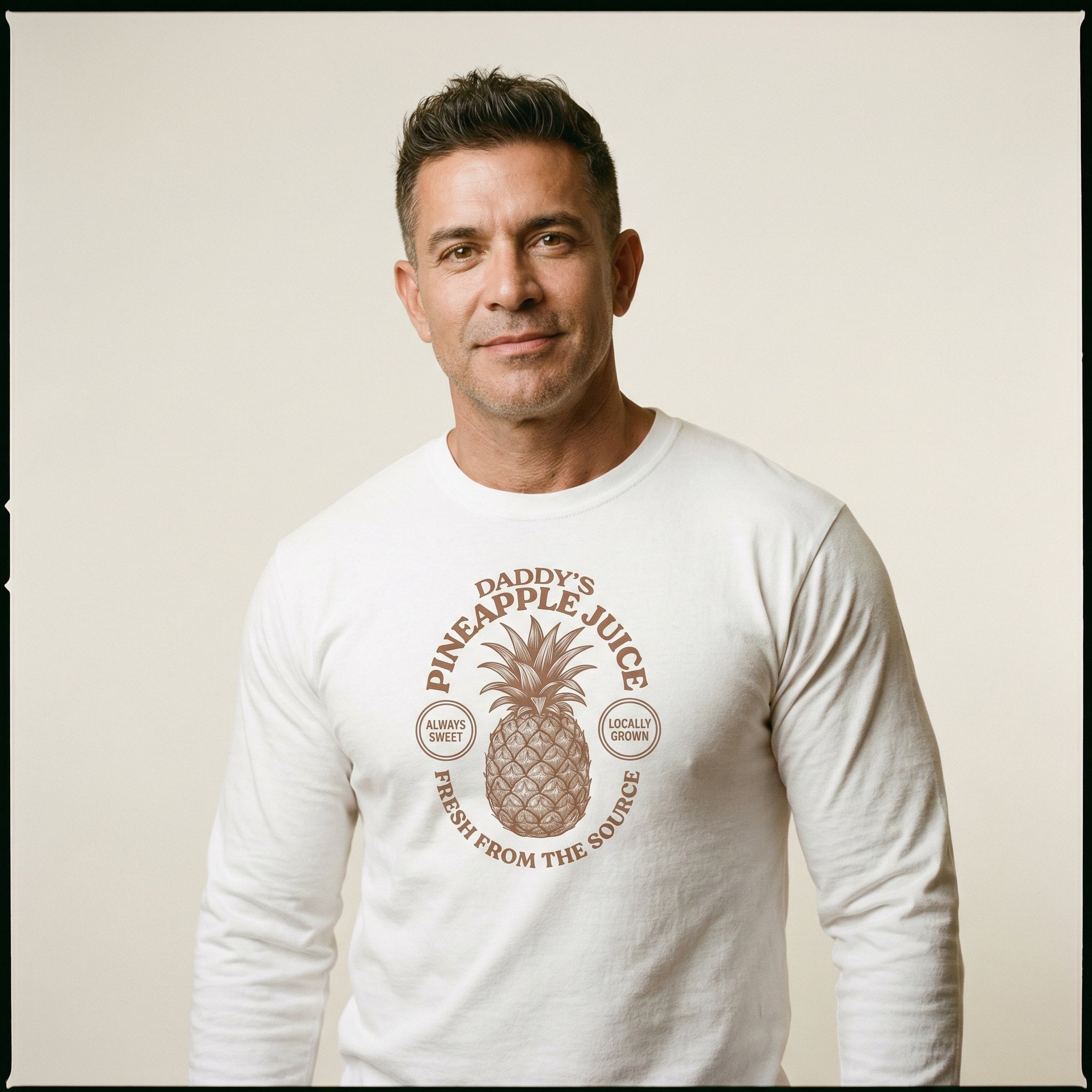 A studio portrait of a middle-aged man with short dark hair and a pleasant smile. He is wearing a white long-sleeve shirt with a central brown graphic. The graphic depicts a pineapple surrounded by the words 'DADDY'S PINEAPPLE JUICE' and 'FRESH FROM THE SOURCE'. Small circular stamps on either side of the pineapple read 'ALWAYS SWEET' and 'LOCALLY GROWN'. The man is posed against a plain, light beige background. #color_White