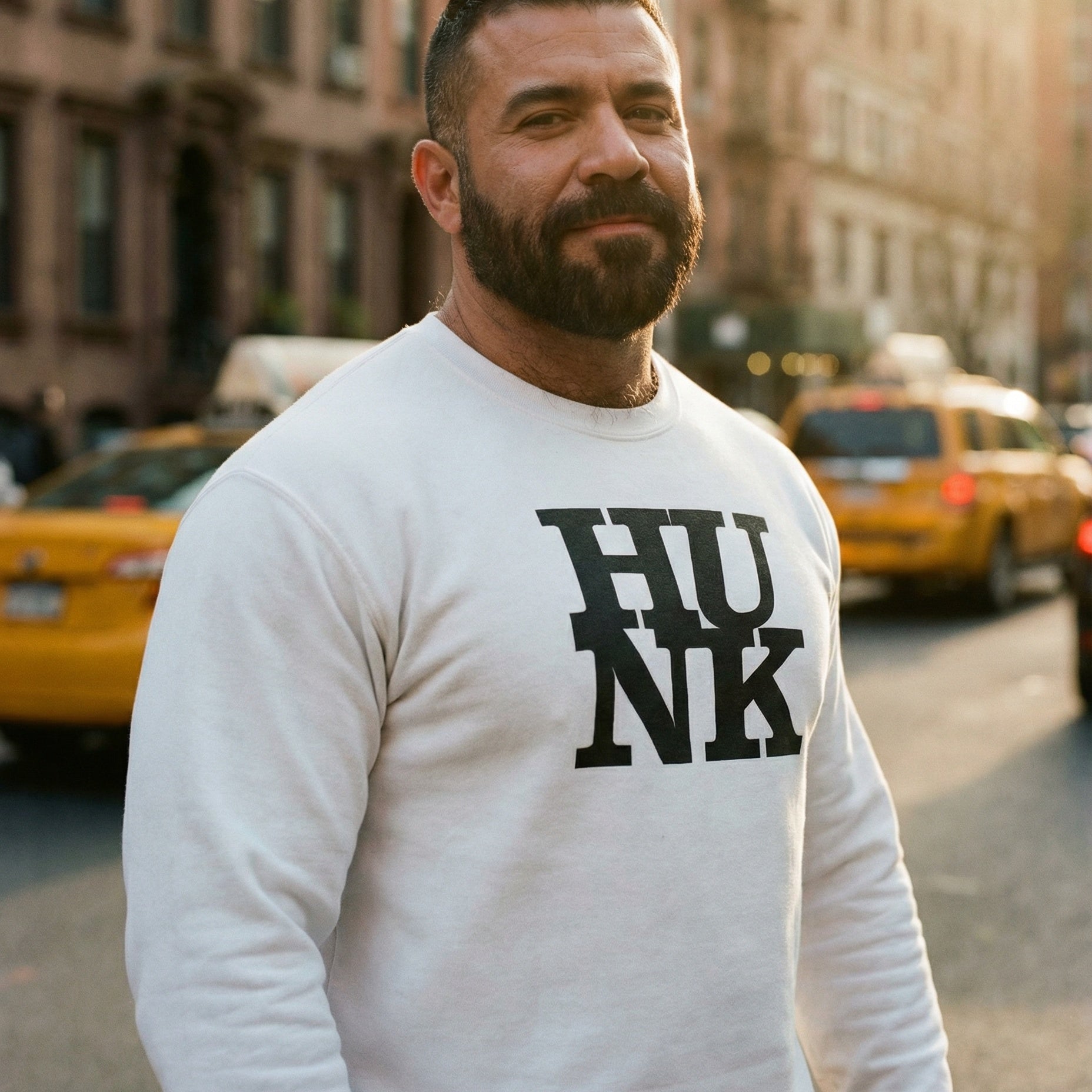 A portrait of a bearded man with short dark hair, standing in the middle of a city street during golden hour. He is wearing a white sweatshirt that has the word 'HUNK' printed in large, bold, black serif font on the chest. The background shows blurred yellow taxi cabs and brownstone buildings under a warm, glowing afternoon sun. The man is looking directly at the camera with a faint, confident smile. #color_White