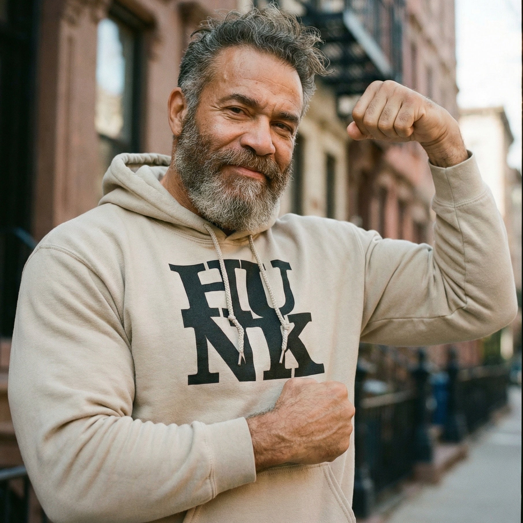 A medium close-up of a middle-aged man with a thick salt-and-pepper beard and messy hair, smiling confidently while flexing his left bicep. He is wearing a tan drawstring hoodie with the word 'FUNK' printed in large, black, stylized letters across the chest. The background shows a sunny, out-of-focus city street lined with brownstone buildings. His right hand is clenched in a fist near his chest, accentuating his strong physique. #color_Sand