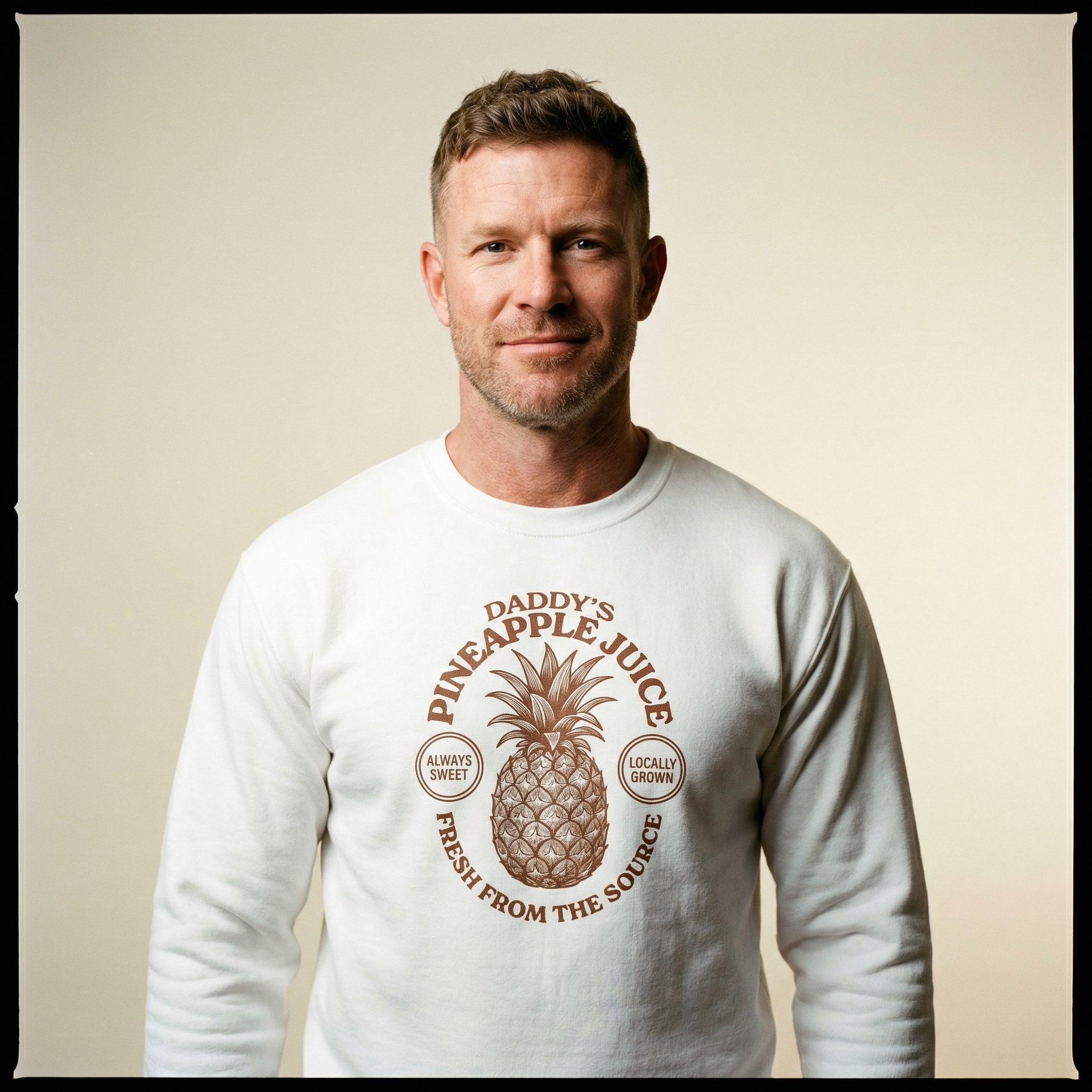 A middle-aged man with short brown hair and light stubble smiles at the camera, wearing a white sweatshirt. The shirt features a large brown graphic of a pineapple with the text 'DADDY'S PINEAPPLE JUICE', 'ALWAYS SWEET', 'LOCALLY GROWN', and 'FRESH FROM THE SOURCE'. He is posed against a plain, neutral background in a studio-style portrait. #color_White