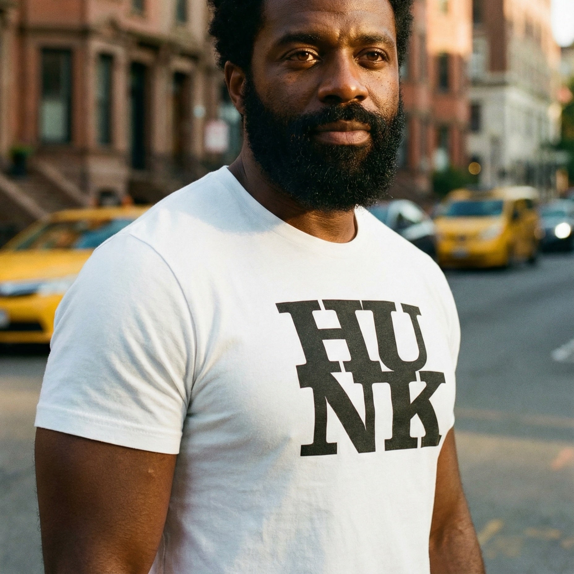 A medium shot of a Black man with a full beard and short dark hair, wearing a white t-shirt that says 'HUNK' in large black letters. He stands on a city street with blurred yellow taxis and brownstone buildings in the background under warm evening light. #color_White