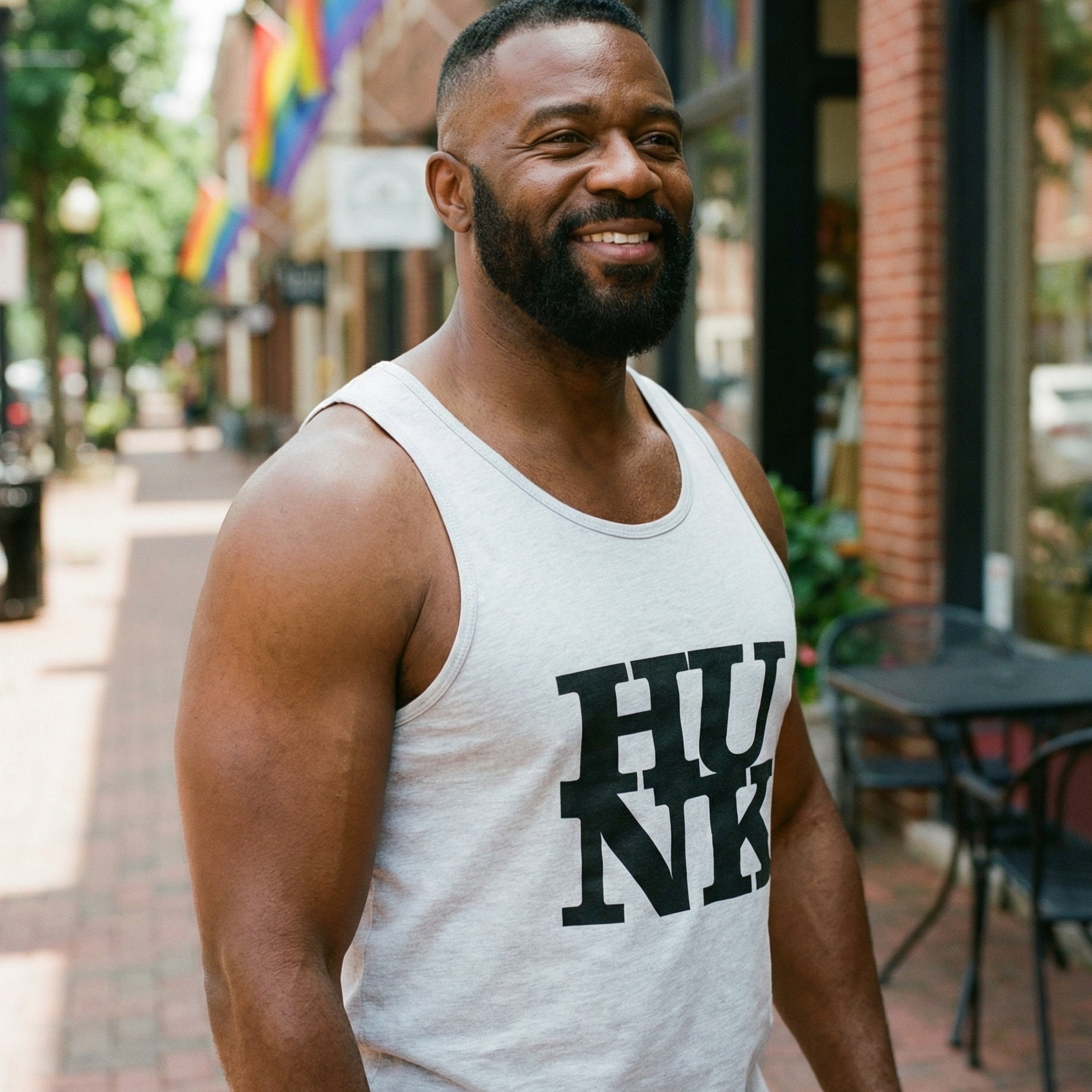 A medium shot of a muscular Black man with a beard and short hair, smiling and looking away from the camera. He is wearing a light gray tank top with the word 'HUNK' in large black block letters. He stands on a brick sidewalk lined with shops, and several rainbow Pride flags hang from the buildings in the background. #color_White